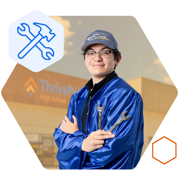 A student is wearing a graduation gown and cap while holding two wrenches and smiling in front of a school building